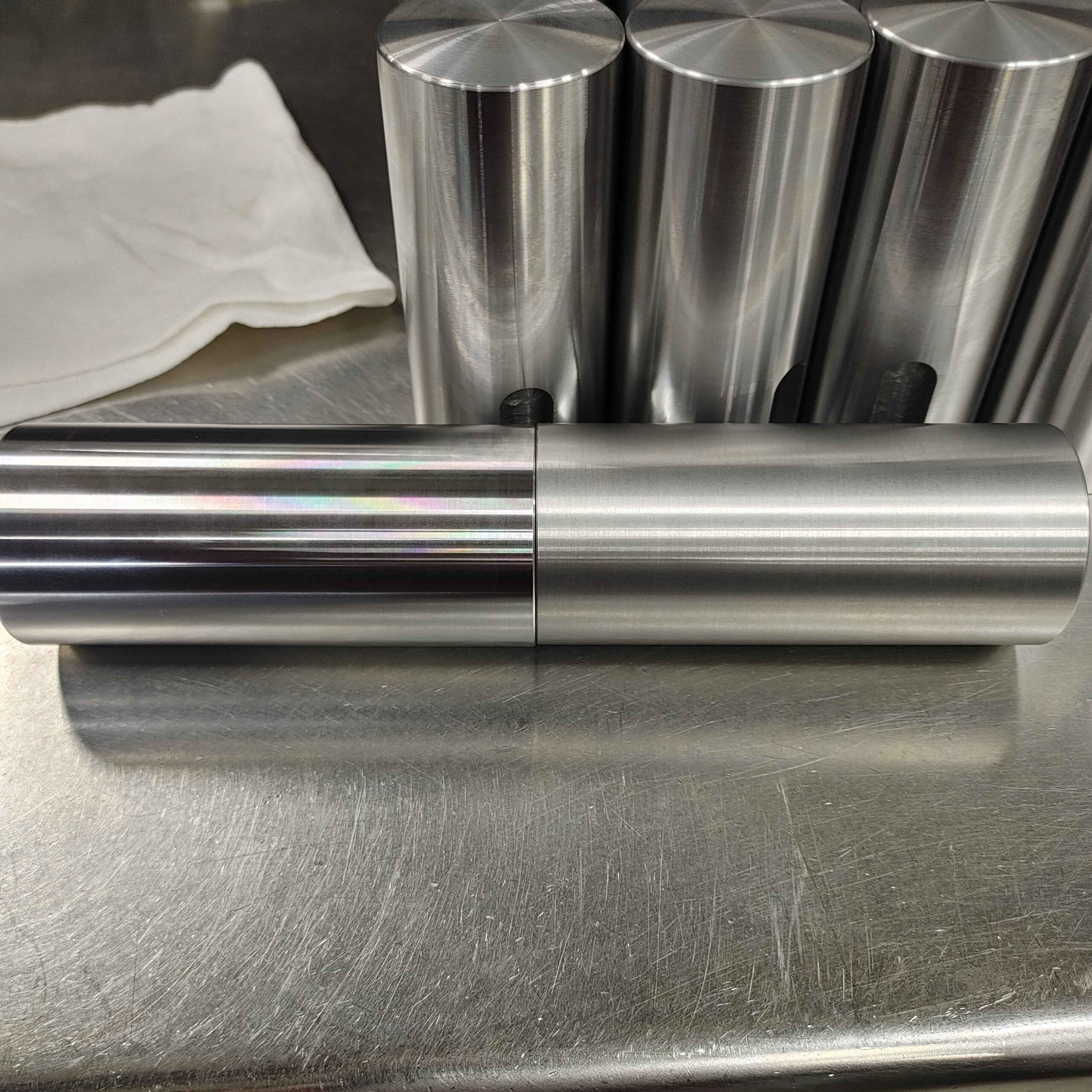A set of cylindrical metallic objects with a brushed finish, likely precision parts or tools, on a metal surface with a cloth in the background.