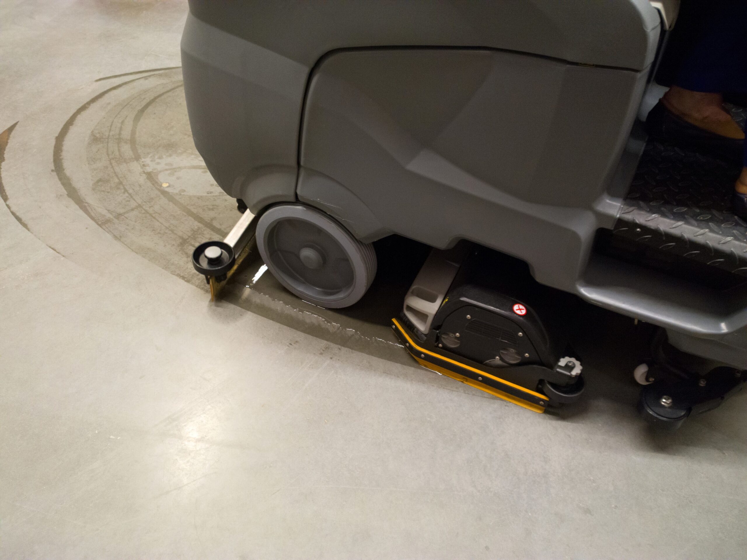 Part of an industrial floor cleaning machine in operation, leaving a wet trail on a concrete floor.