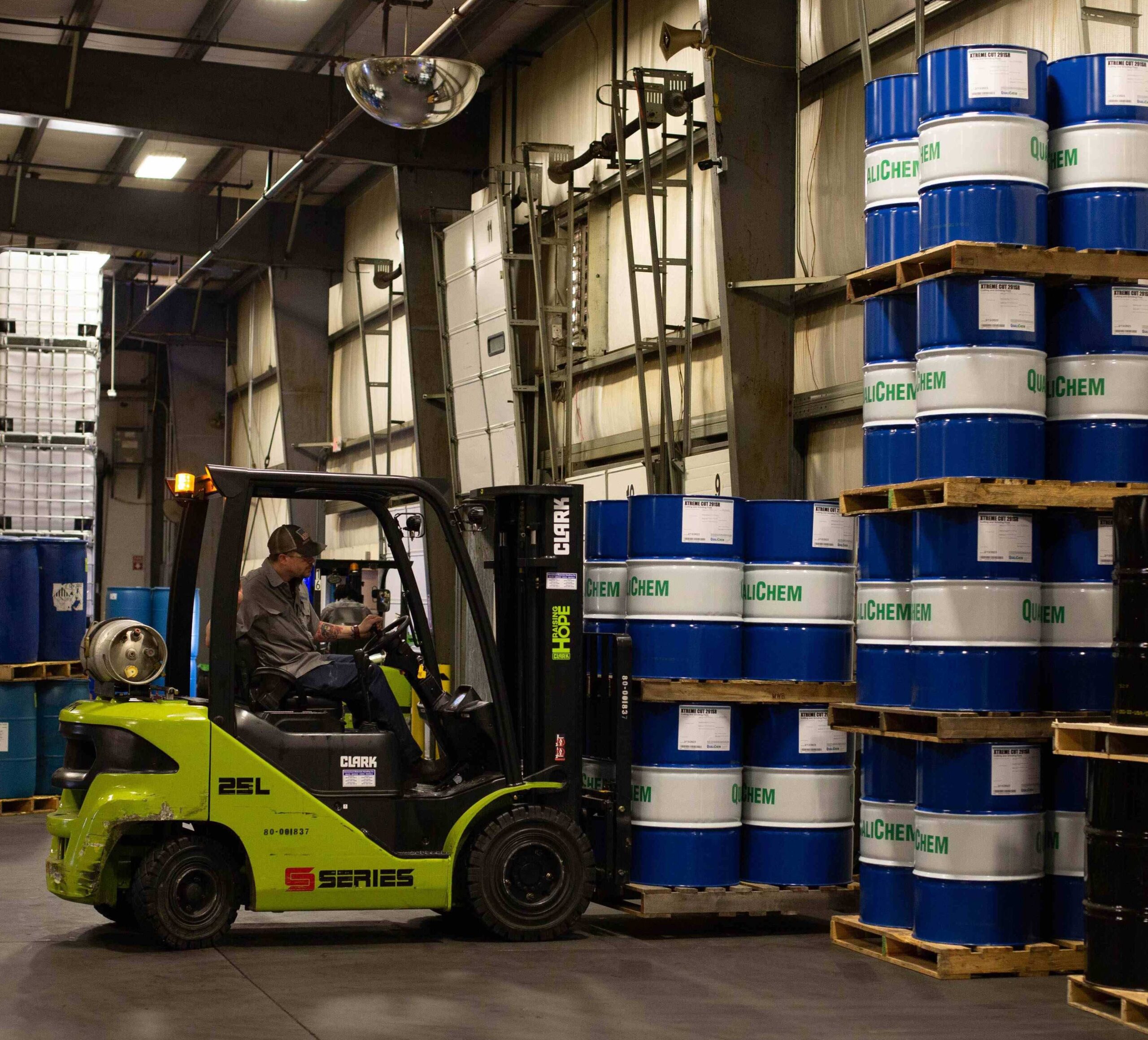 A forklift operator transporting blue chemical drums inside a warehouse with shelves stocked with various industrial containers.