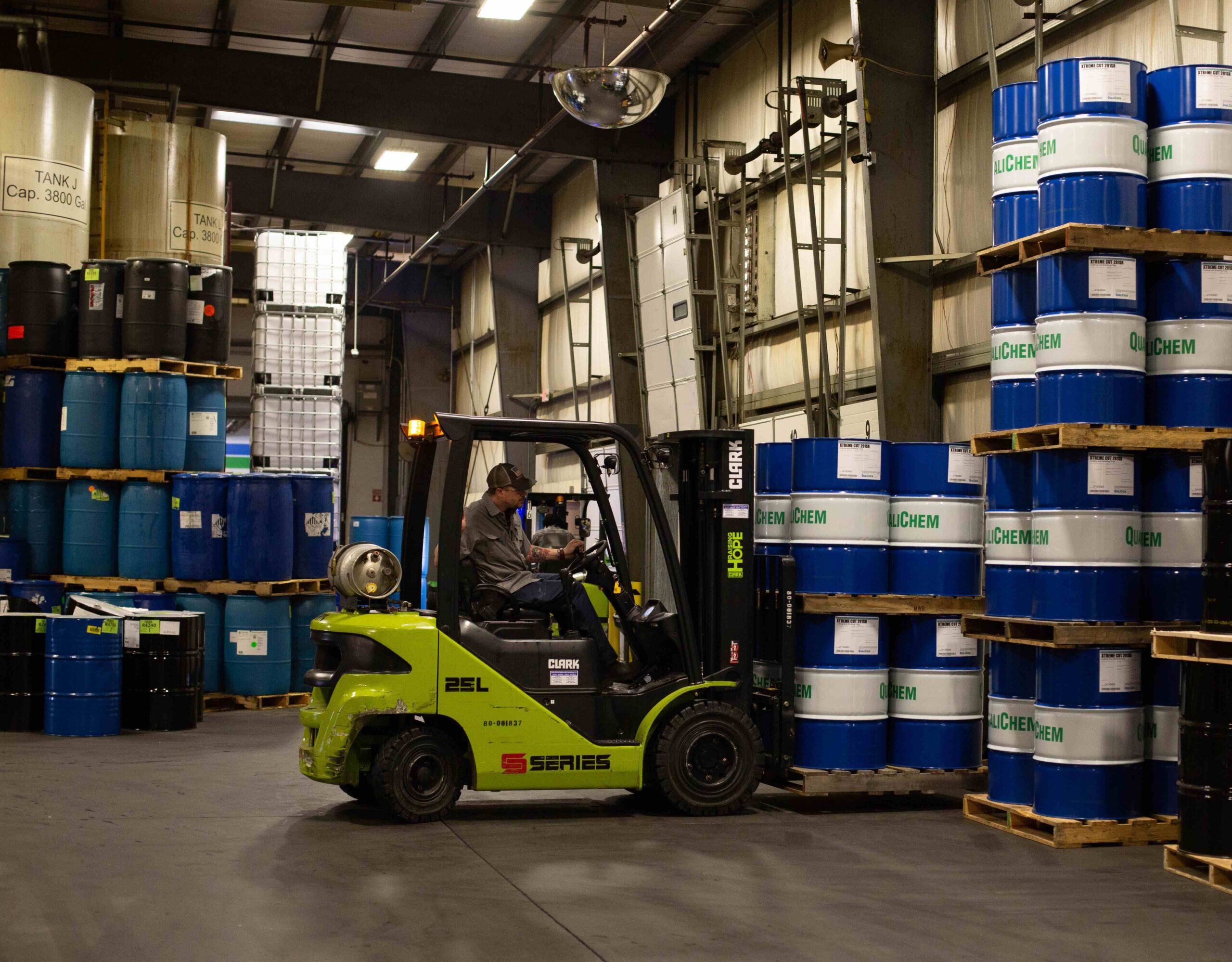 A forklift operator transporting blue chemical drums inside a warehouse with shelves stocked with various industrial containers.