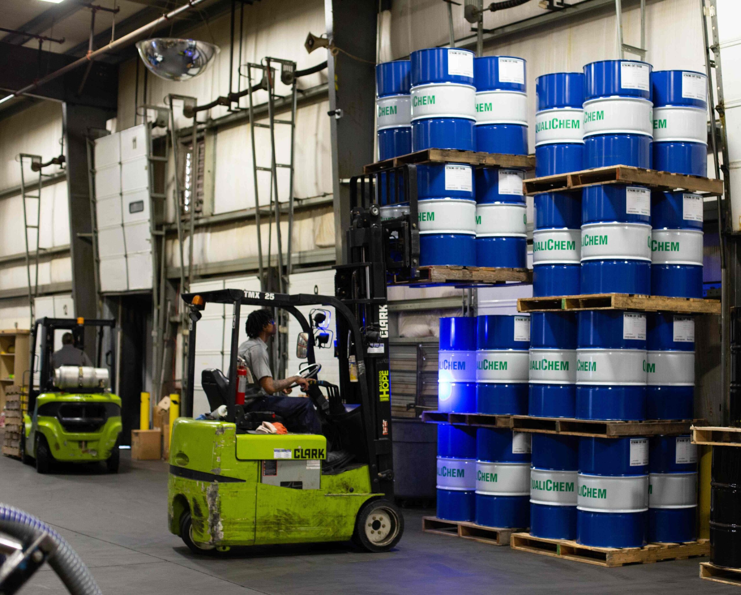A forklift operator transporting blue chemical drums inside a warehouse with shelves stocked with various industrial containers.