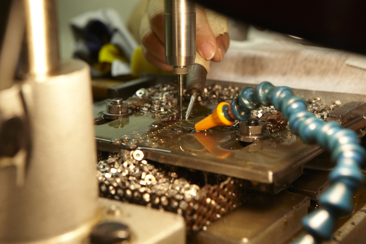 Close-up of a drilling process on a metal part with metal shavings around, and a cooling hose directing fluid onto the work area.