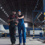 Two workers in a large industrial hangar; one in a black cap and red checked shirt holding a tablet, and the other in a blue jumpsuit, both looking at the tablet screen.